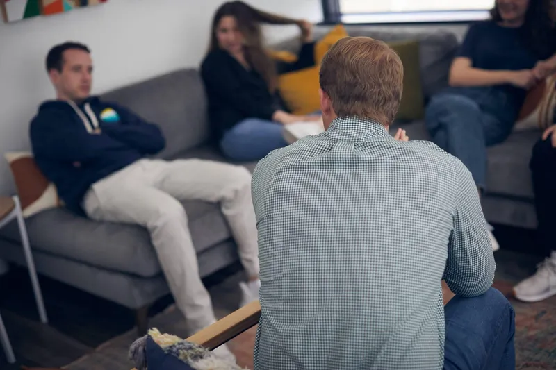 A man seen from behind in a checked shirt sitting across from adults on gray couches during a group therapy session