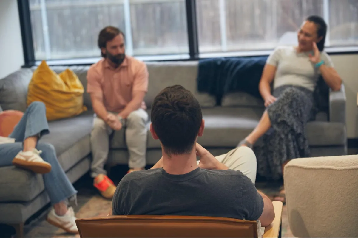 Adults sitting on gray couches by a large window, talking during a group counseling session