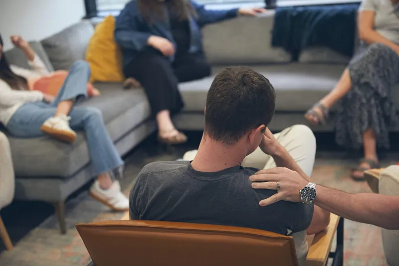 A man seen from behind in a group therapy session as another person rests a supportive hand on his shoulder