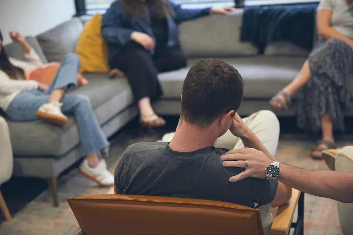 A man seen from behind in a group therapy session as another person rests a supportive hand on his shoulder