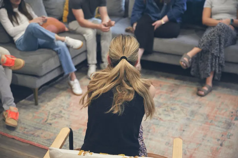 A woman with a blonde ponytail seen from behind sitting in a chair facing adults on gray couches during a group therapy session