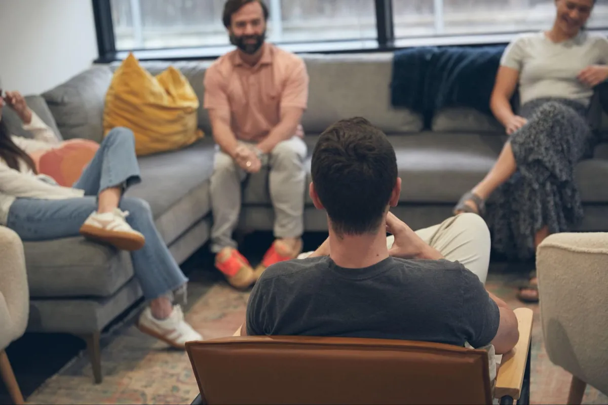 A man seen from behind sitting in a chair across from adults on gray couches during a group therapy session