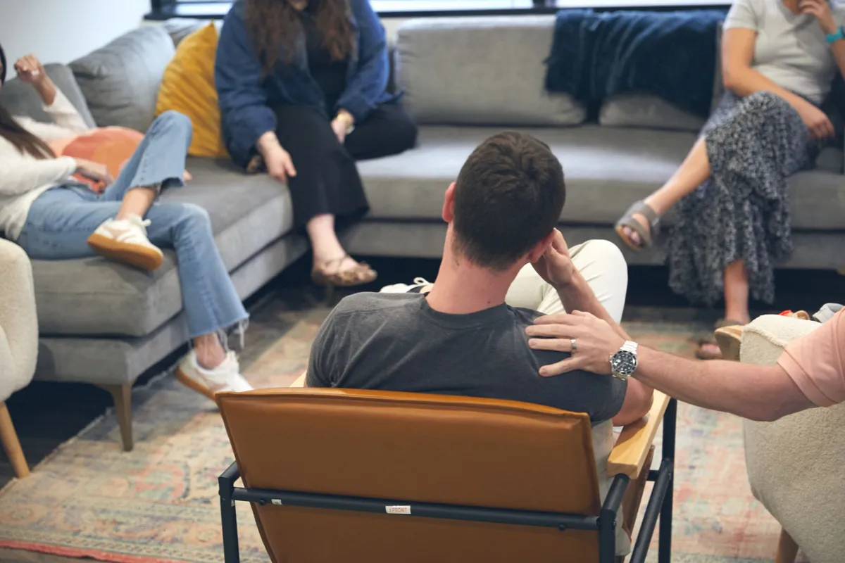 A young man seen from behind in a leather chair during a residential rehab group session as a peer places a supportive hand on his shoulder