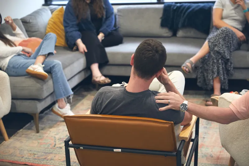 A young man seen from behind in a leather chair during a residential rehab group session as a peer places a supportive hand on his shoulder