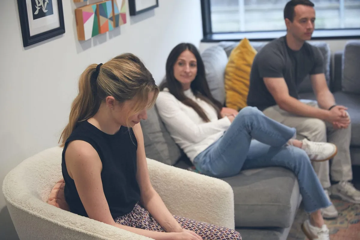 A young woman looking down in an armchair while two adults on a gray couch listen during a group therapy session