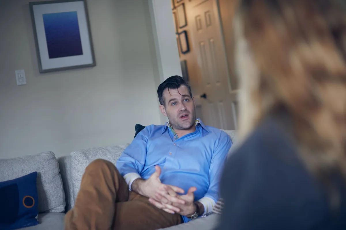 A man in a blue shirt sitting on a gray couch and talking with a therapist during an individual counseling session