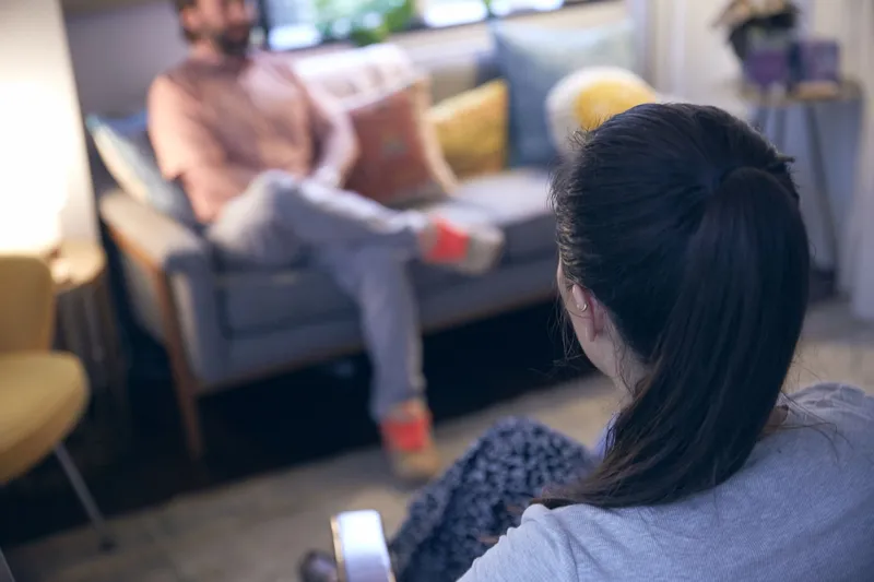 A woman with a dark ponytail seen from behind sitting across from a man on a couch during a mental health assessment